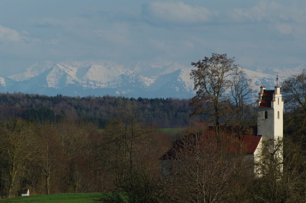 Landschaft mit Kirche im Vordergrund, Wald und weiter Blick auf schneebedeckte Alpen im Hintergrund