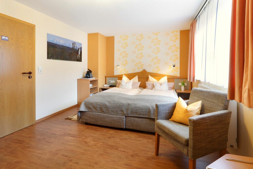 Helles Komfort-Doppelzimmer mit großem Bett, Sitzsessel, warmen Farbtönen und großem Fenster